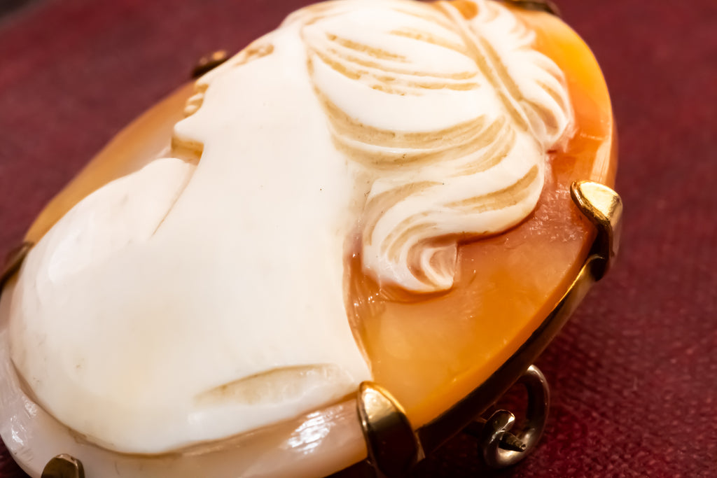 A close-up of a carved oval cameo-style pendant or brooch featuring a white classical profile and flowing hair over a warm amber-orange base, set in gold-toned metal against a dark red background.