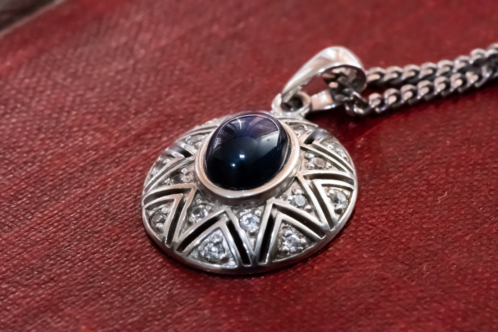 A silver pendant necklace featuring a central dark blue oval gemstone surrounded by intricate silver geometric patterns and small clear stones, resting on a textured red surface.