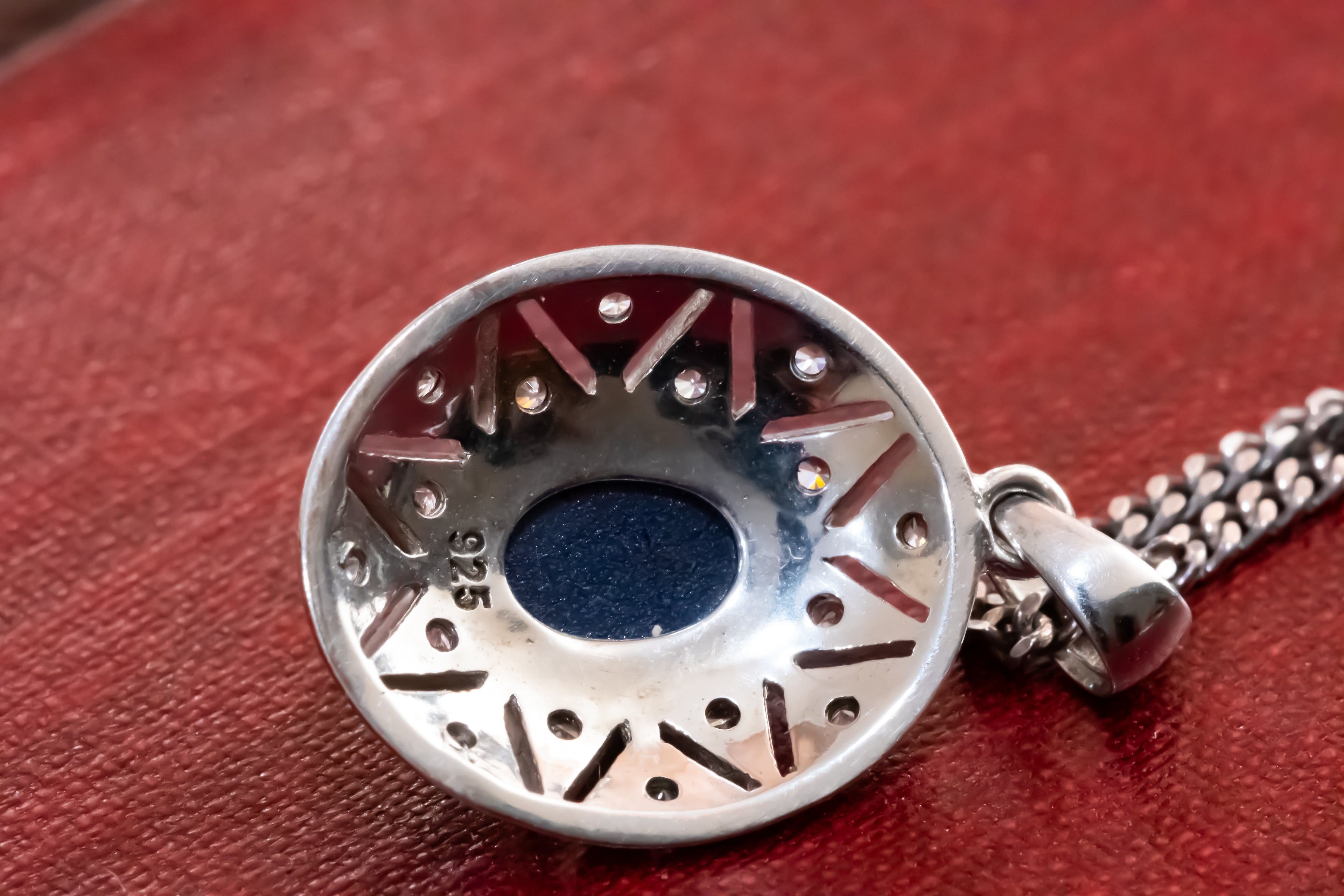 A sterling silver pendant necklace with a circular, perforated design and a central dark blue inlay, resting on a textured red surface.