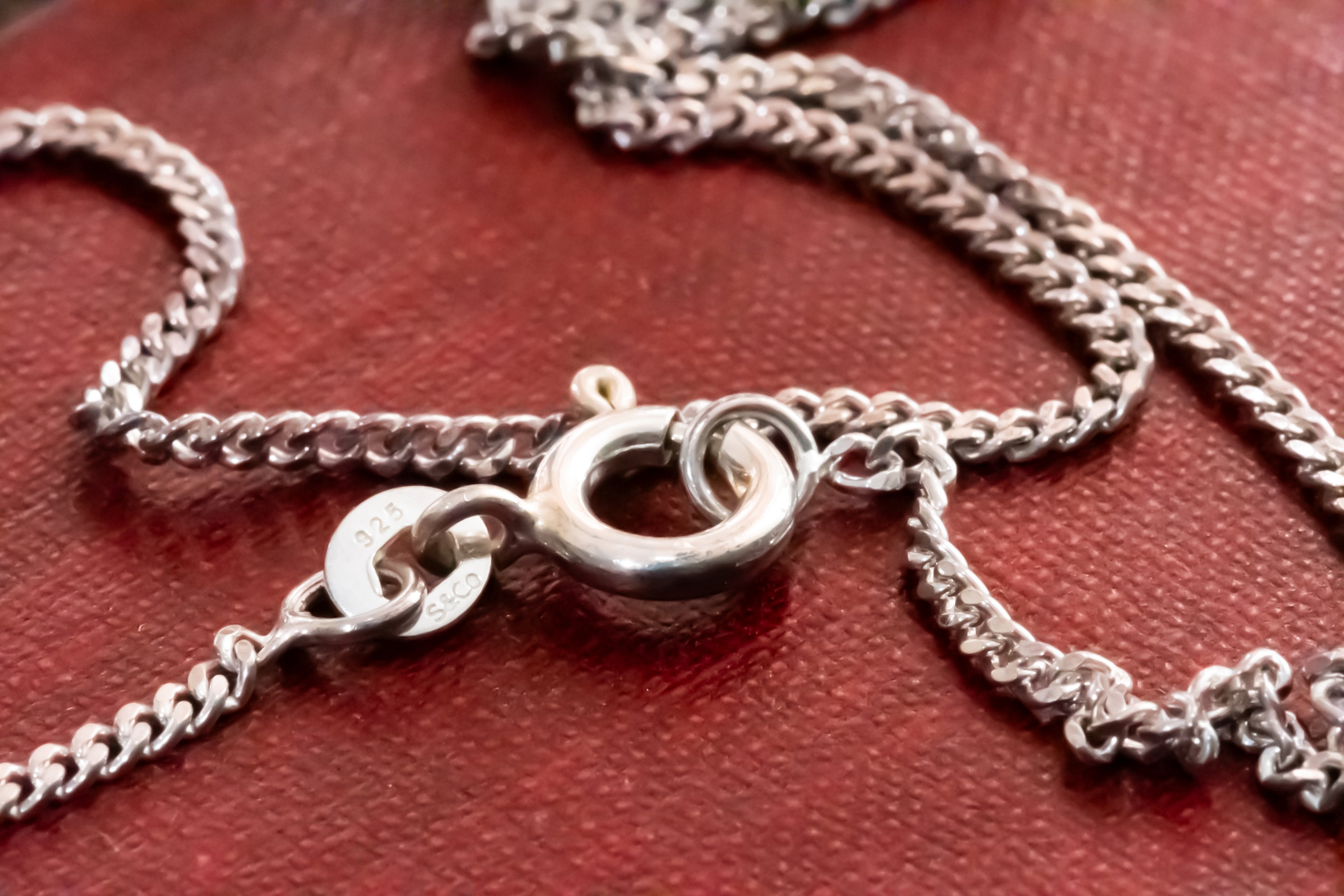 Close-up of a silver chain necklace with a lobster clasp and extension chain, resting on a textured dark red surface.