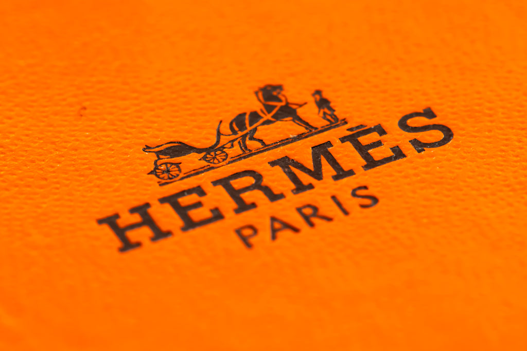 Close-up of a textured bright orange surface with a black Hermès Paris logo and horse-and-carriage emblem printed on it.