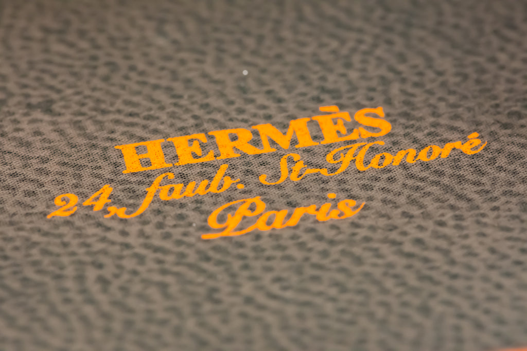 Close-up of textured gray pebbled leather with orange Hermès logo text reading 'HERMÈS 24 Faub. St-Honoré Paris'.