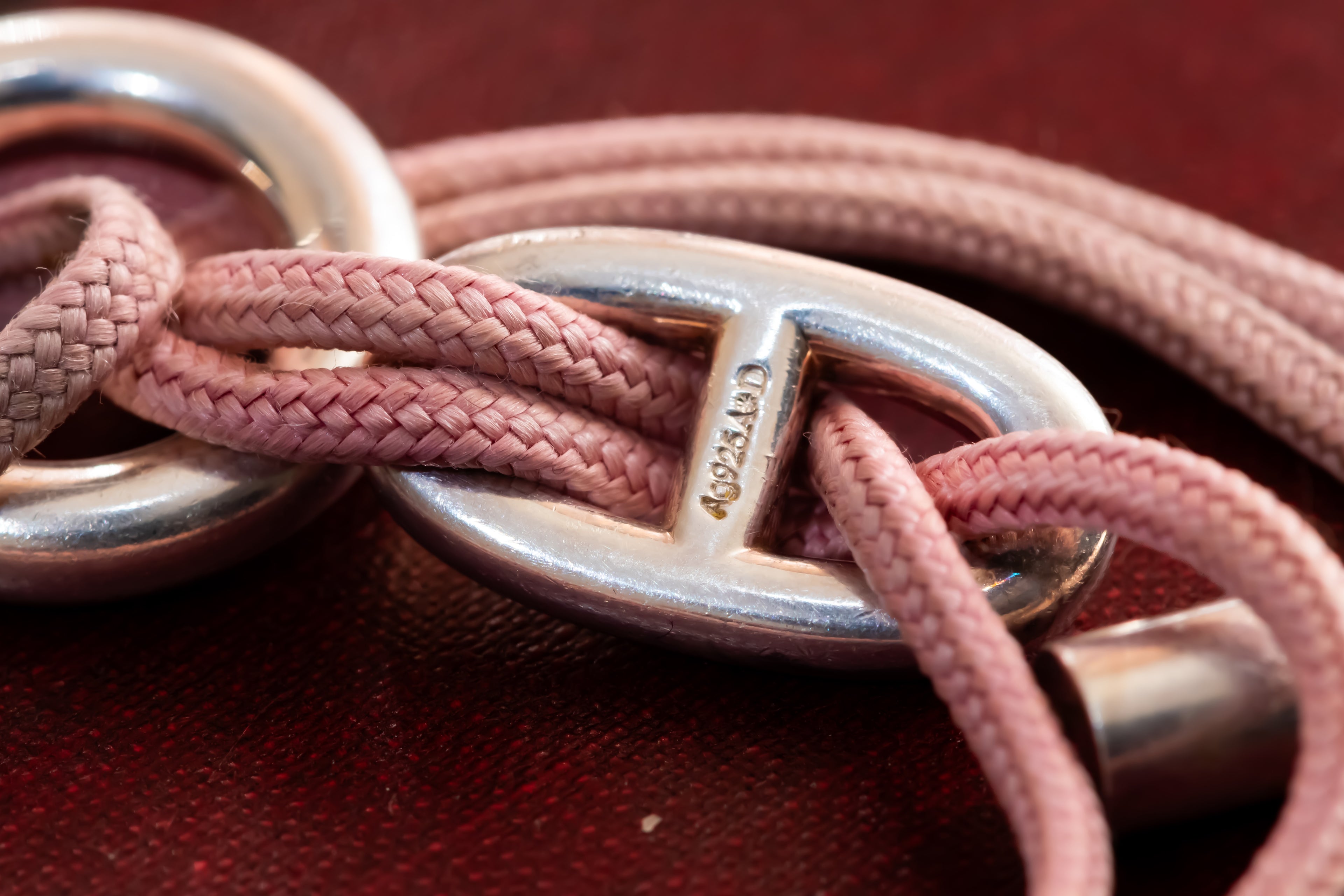 A close-up of a pink braided leather strap threaded through polished silver-tone metal rings and oval links, with a textured dark red surface in the background.