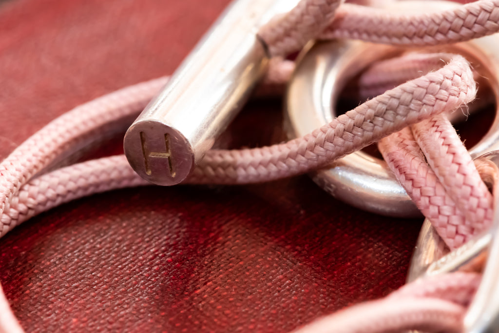 A close-up of a pink braided rope accessory with a polished silver metal rod engraved with the letter 'H', resting on a textured dark red surface.