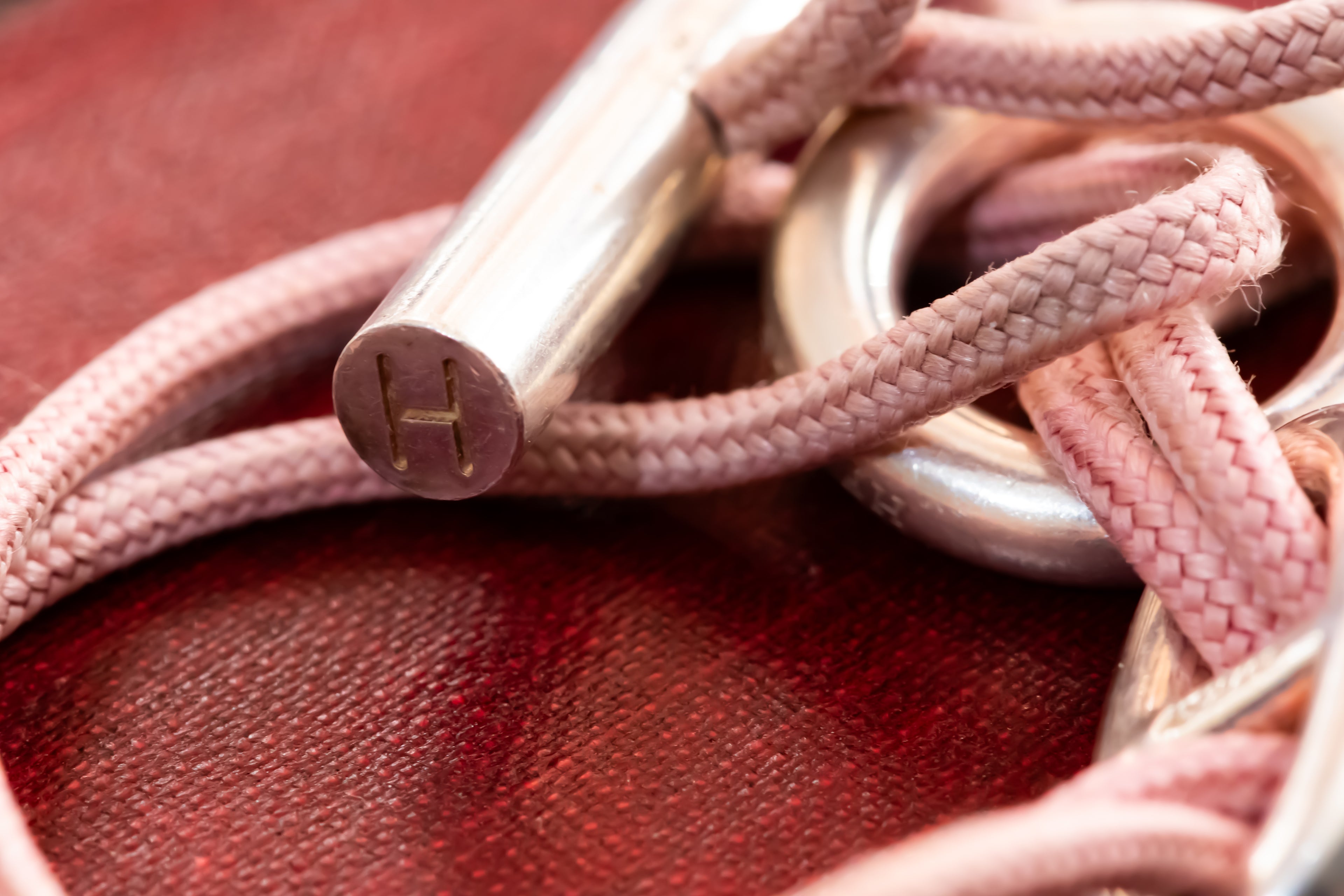 A close-up of a pink braided rope accessory with a polished silver metal rod engraved with the letter 'H', resting on a textured dark red surface.