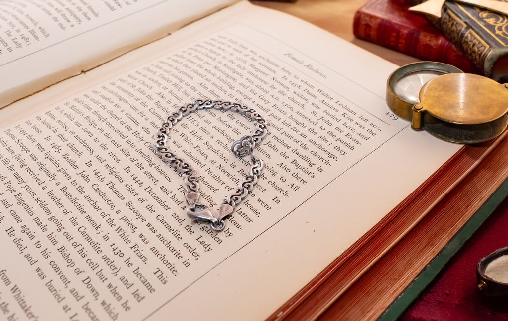 A silver chain bracelet with a lobster clasp and toggle closure rests on the pages of an open vintage book, surrounded by old books and a brass pocket mirror on a wooden surface.