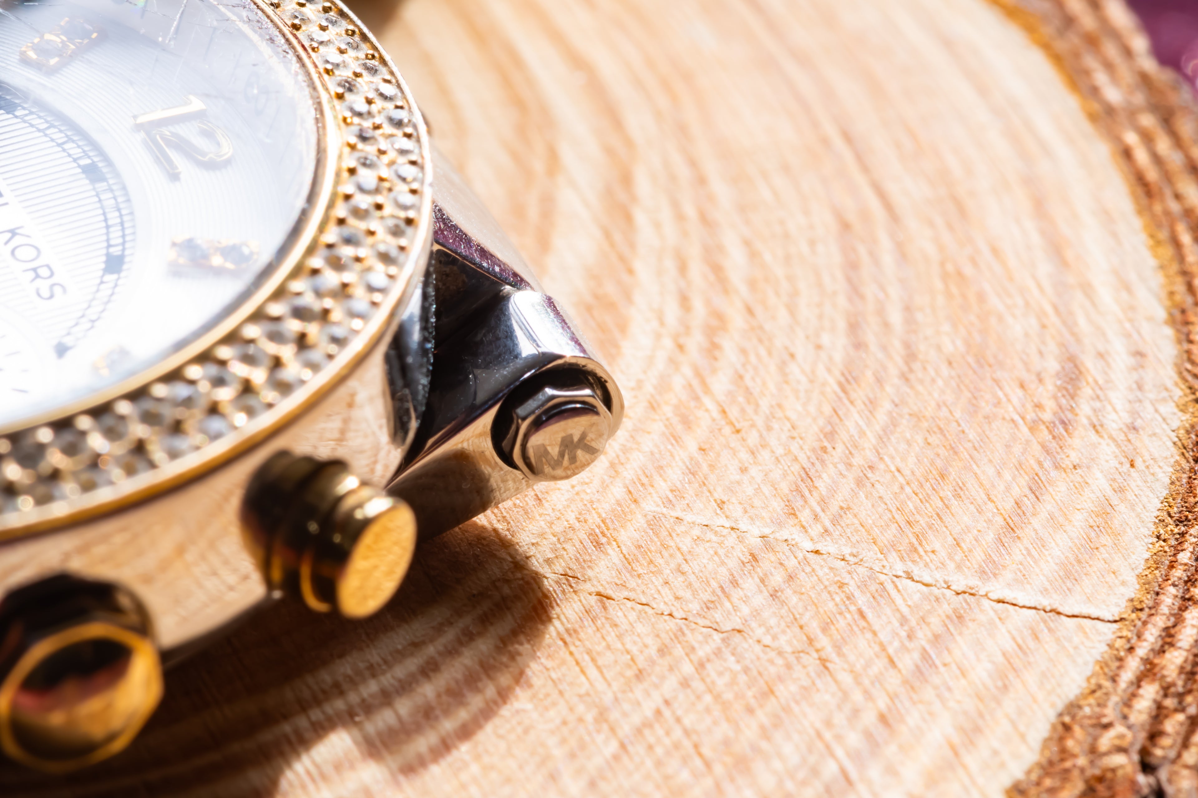 Close-up of a luxury watch with a gold bezel, white dial, and polished silver-toned case, resting on a textured wood slice background.