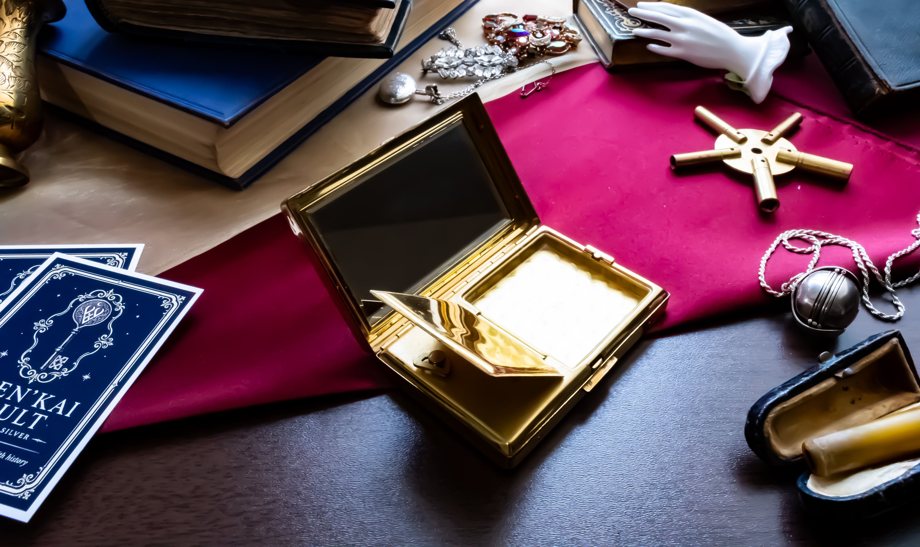 An ornate open gold case with a mirror and gold interior, styled on a dark wooden surface with a red cloth, old books, jewelry, and a blue card in a moody tabletop arrangement.