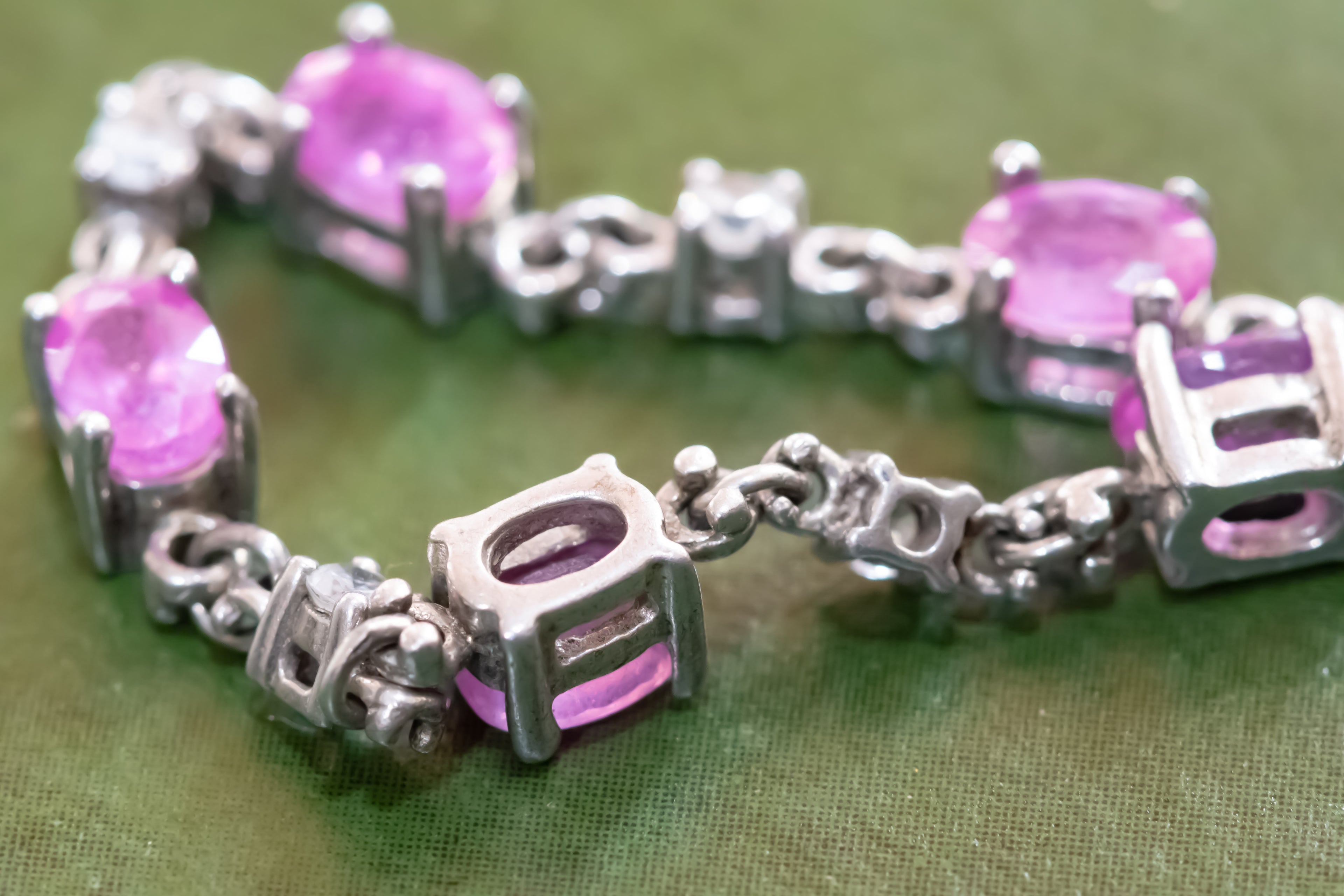 A close-up of a silver-toned bracelet featuring oval pink gemstones and decorative cube-shaped links, displayed on a textured olive green fabric background.
