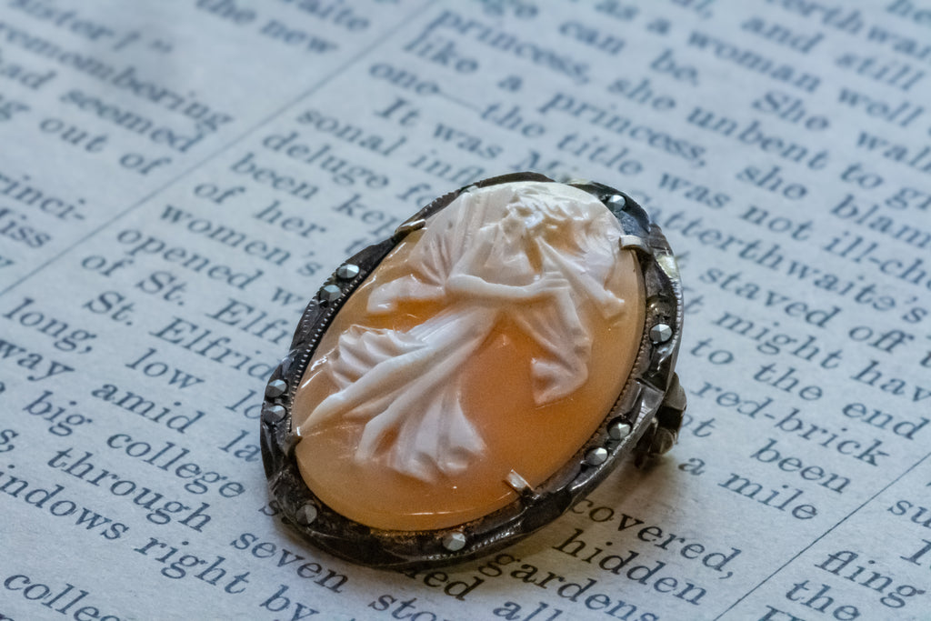 A vintage-style carved cameo featuring a white figure relief on a warm amber-orange background, set in a dark silver-toned metal bezel with small marcasites, resting on printed book text.