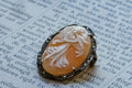 A vintage-style carved cameo featuring a white figure relief on a warm amber-orange background, set in a dark silver-toned metal bezel with small marcasites, resting on printed book text.