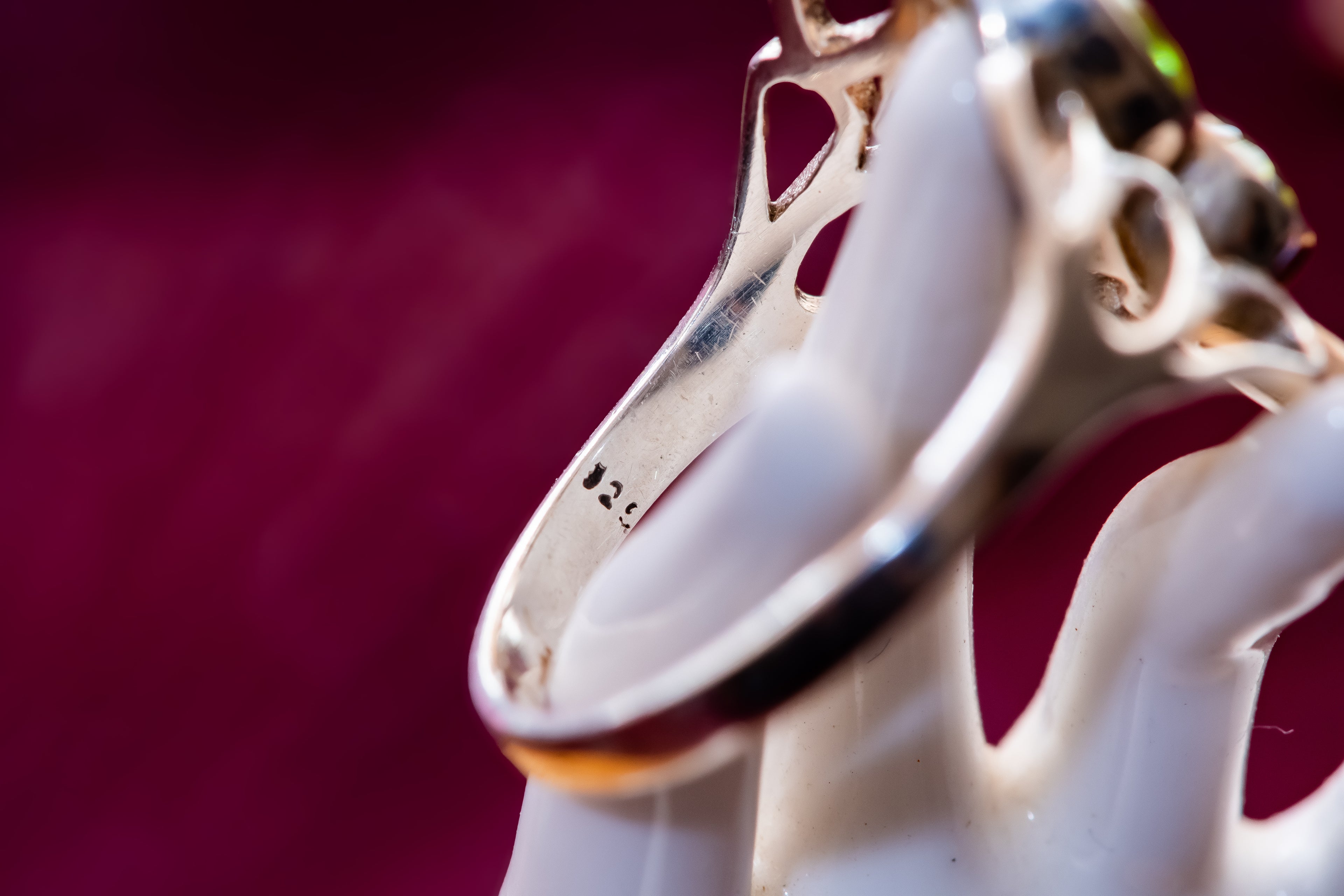 A close-up shot of a silver ring with a decorative filigree band, resting on a white ceramic ring holder against a dark magenta background. The inner band is stamped with '925'.