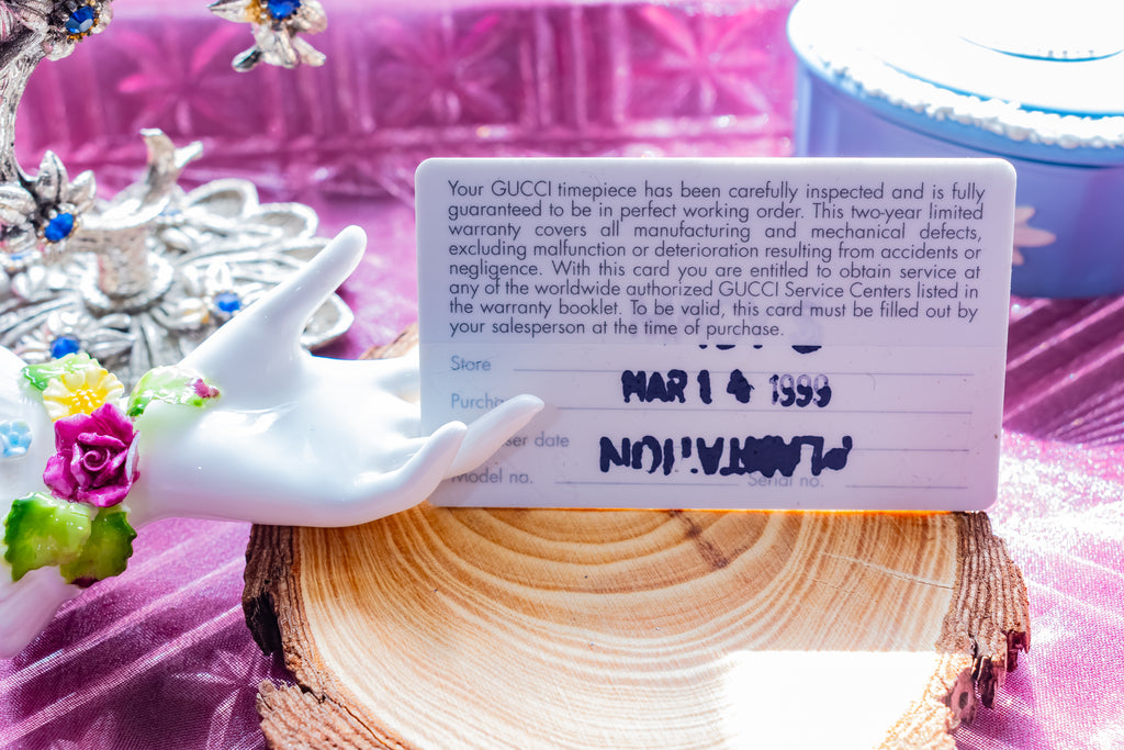 A GUCCI limited warranty card displayed on a wooden slice stand, surrounded by decorative props including a white hand figurine, flowers, and a purple fabric background.