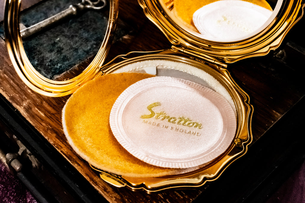 An open gold-framed Stratton compact case with a white powder puff and gold logo, resting on a dark wooden surface.
