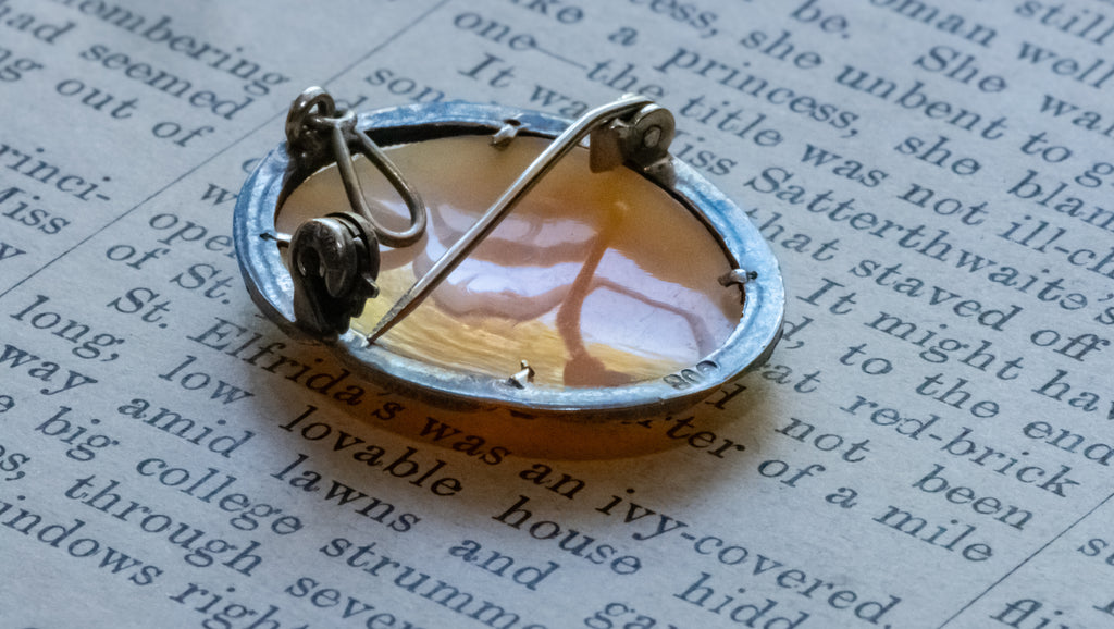 A vintage-style oval brooch with a silver-toned metal frame and a warm amber-orange translucent center, photographed resting on printed book pages.