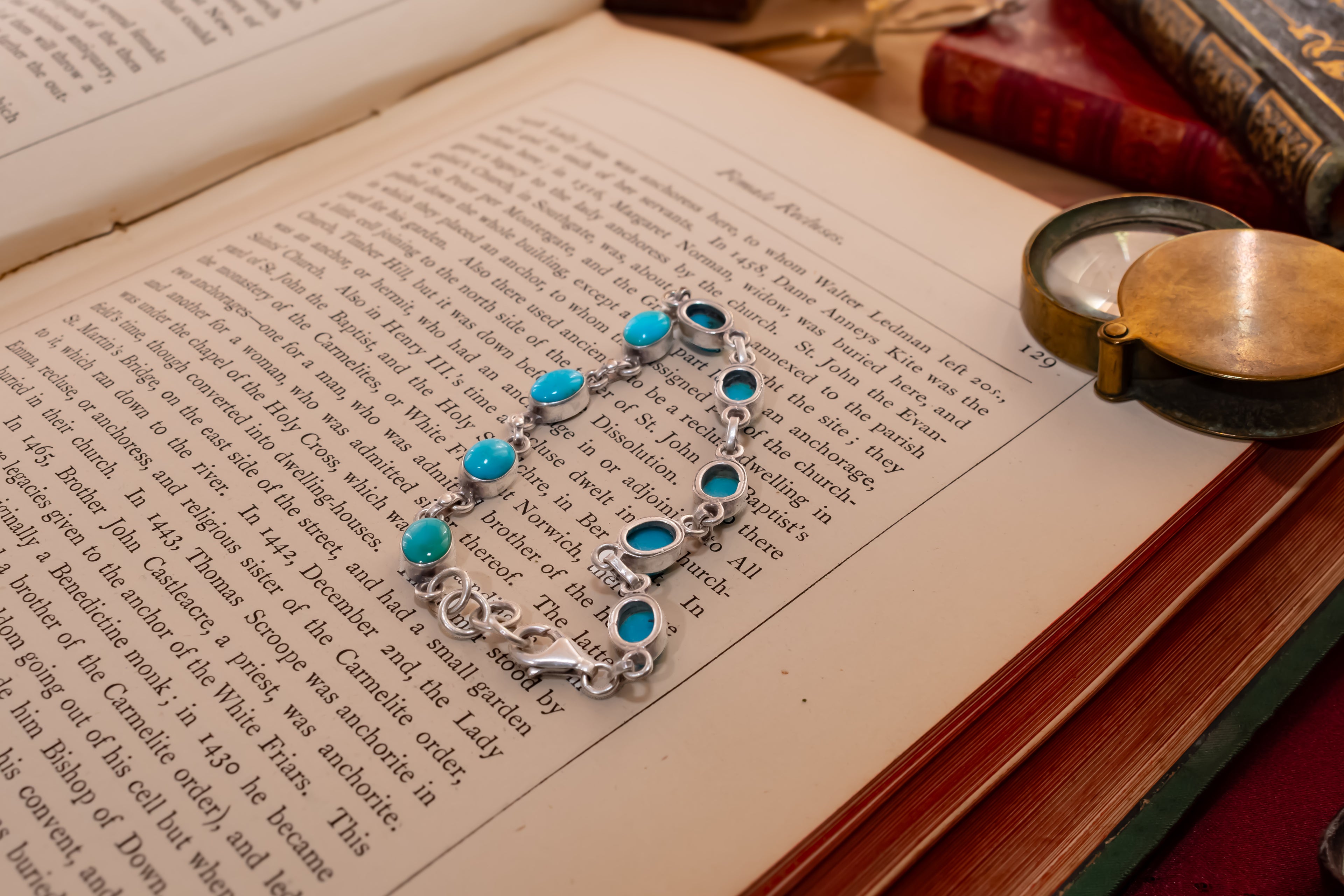 A silver-toned chain bracelet with multiple oval turquoise-blue stones, laid across the pages of an open book. The scene includes vintage books and a brass pocket mirror on a wooden surface.