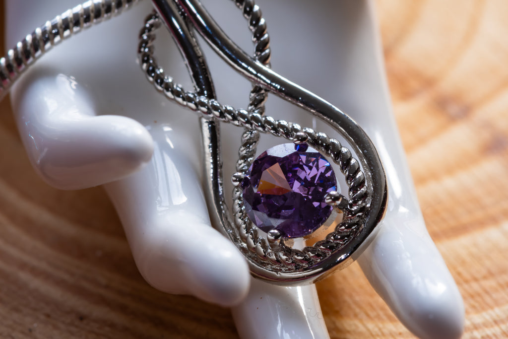 A silver-toned pendant necklace with a twisted rope design and a round faceted purple gemstone, displayed on a white ceramic mannequin hand over a wooden surface.