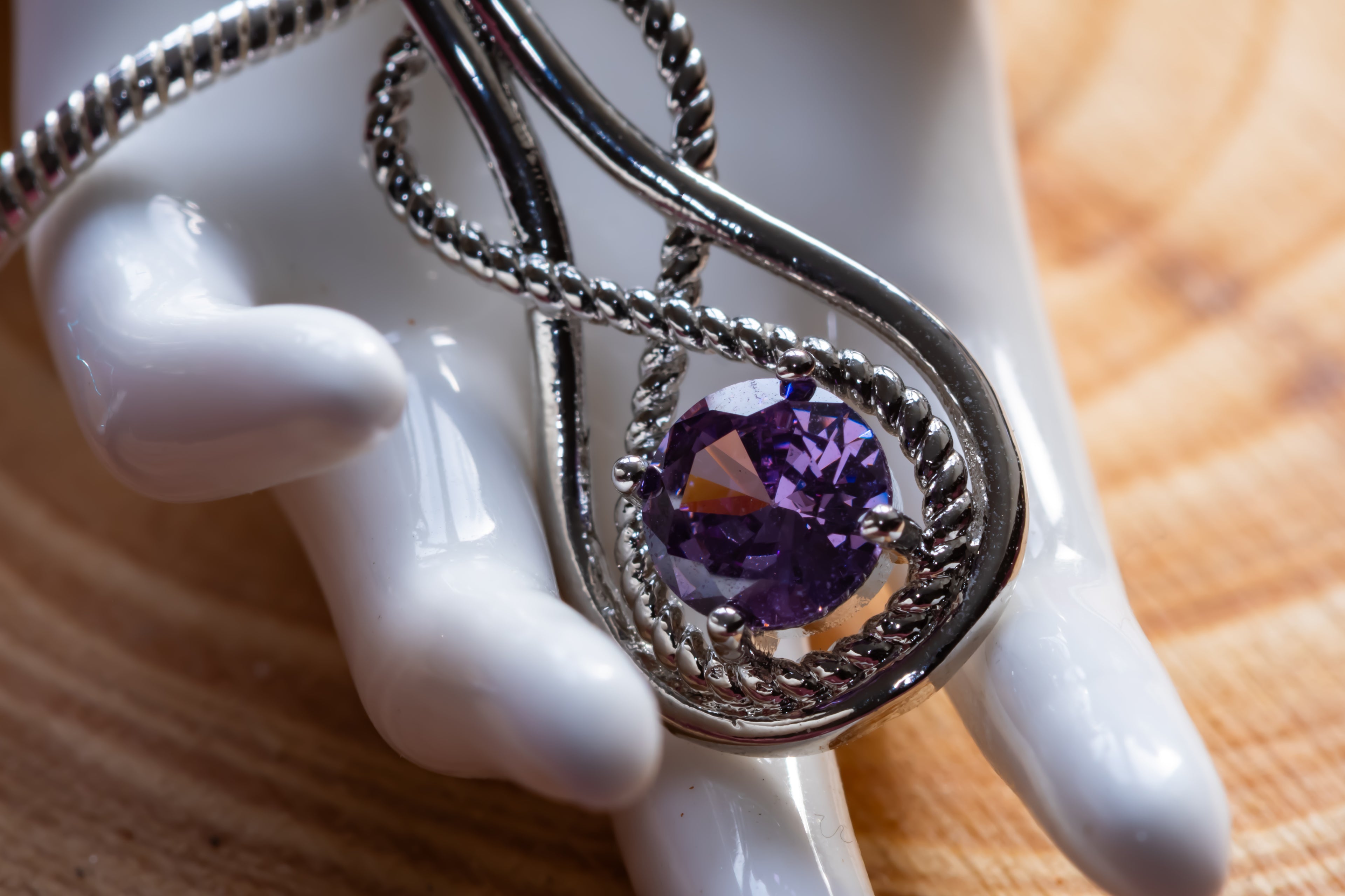 A silver-toned pendant necklace with a twisted rope design and a round faceted purple gemstone, displayed on a white ceramic mannequin hand over a wooden surface.