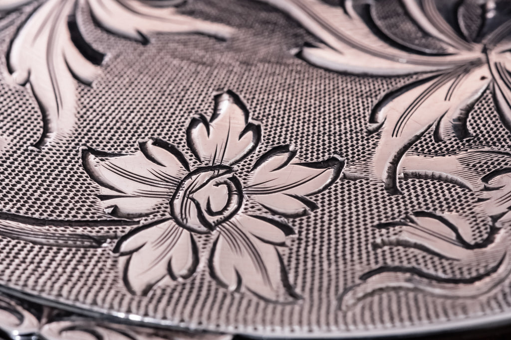 Close-up of an ornate silver powder compact with embossed floral motifs and a textured woven-like pattern, shown in warm silver and rose-gold tones.