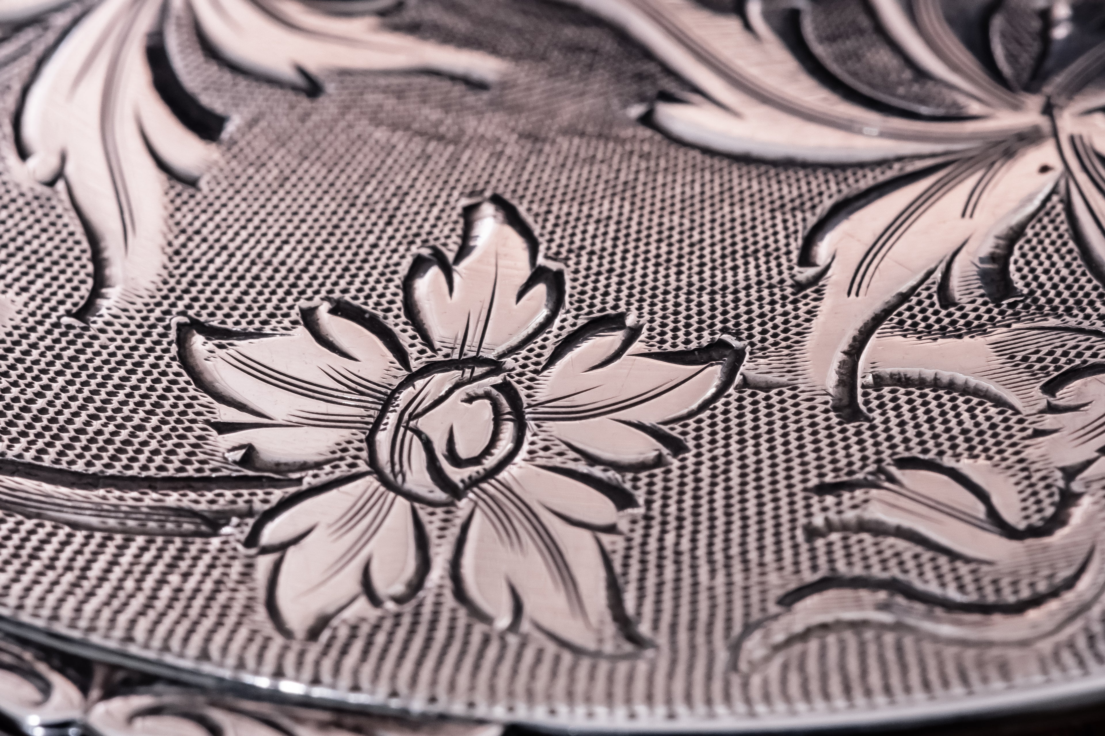 Close-up of an ornate silver powder compact with embossed floral motifs and a textured woven-like pattern, shown in warm silver and rose-gold tones.