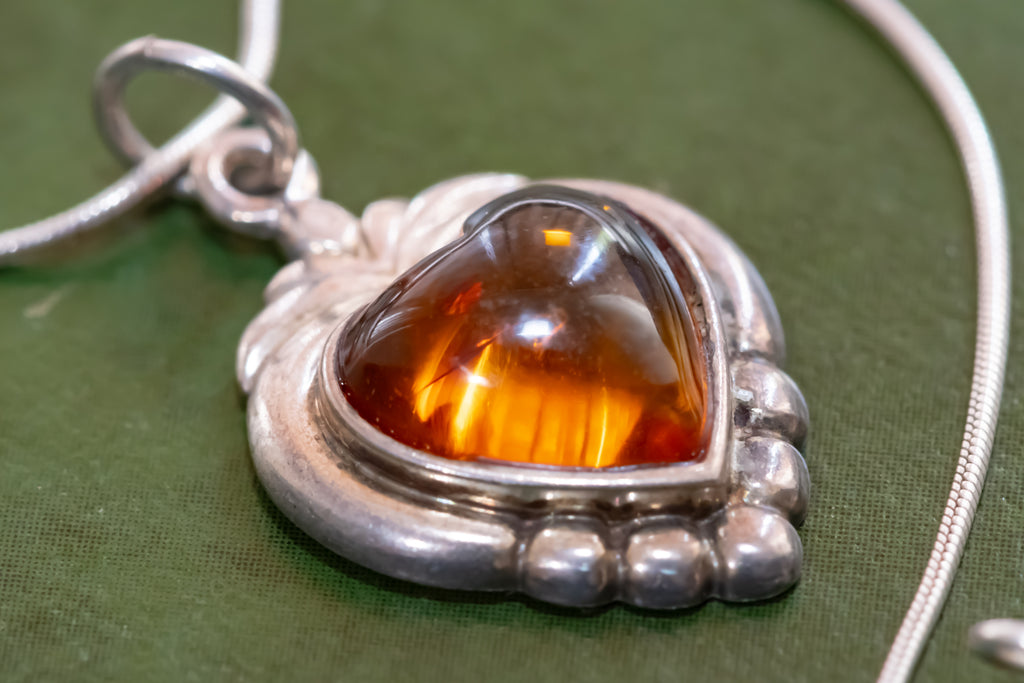 A close-up of a heart-shaped amber pendant in a silver-toned setting with a snake chain, resting on a textured green surface.