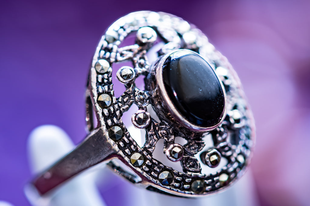 An ornate silver-toned ring with a large oval black cabochon stone and detailed filigree settings, shown close-up against a soft purple background.