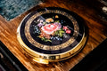 A round compact mirror with a black and gold lid featuring a hand-painted pink rose floral design, resting on a rustic wooden surface.