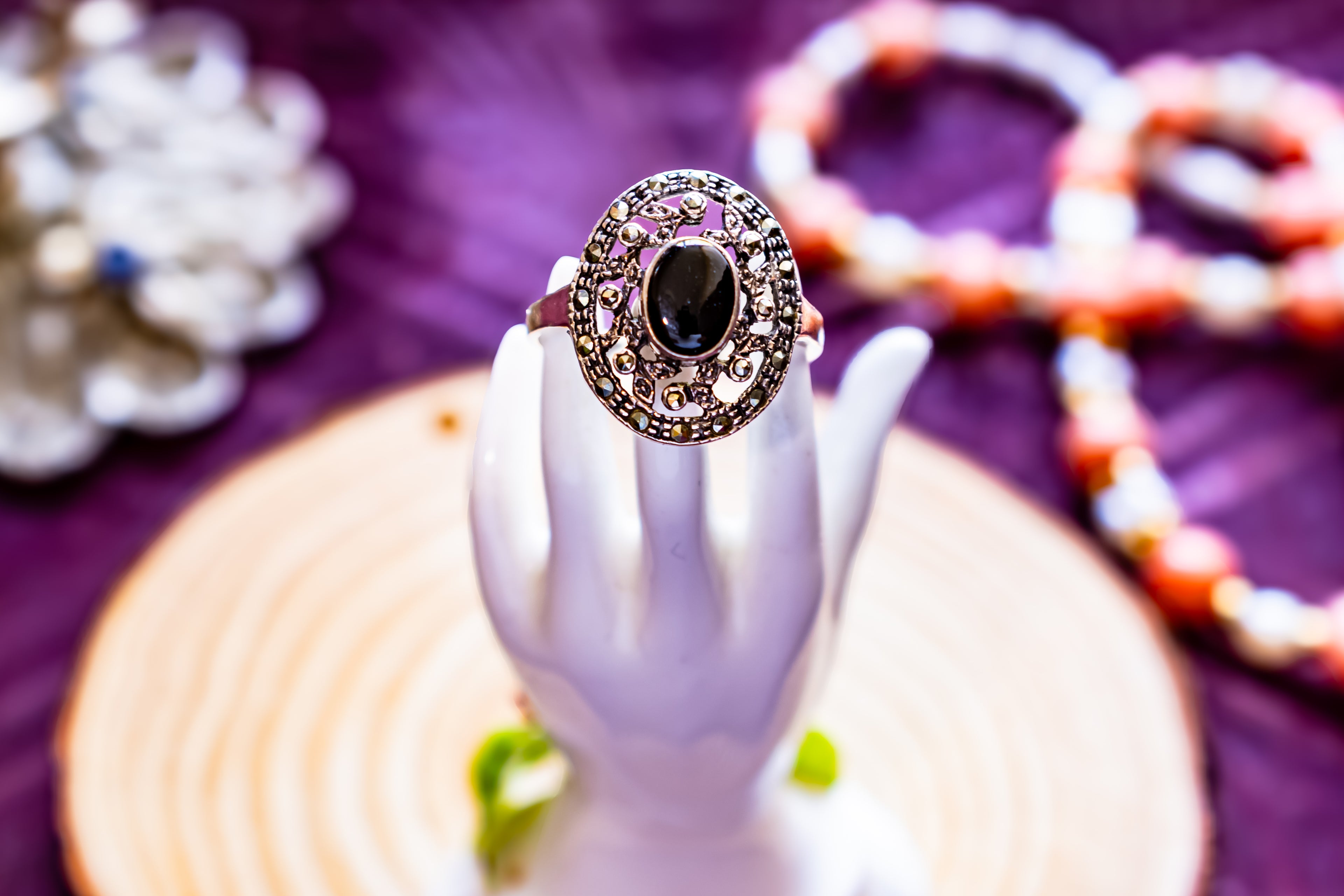 An ornate silver ring with a large oval black gemstone and filigree detailing, displayed on a white hand-shaped stand against a purple background with blurred jewelry.