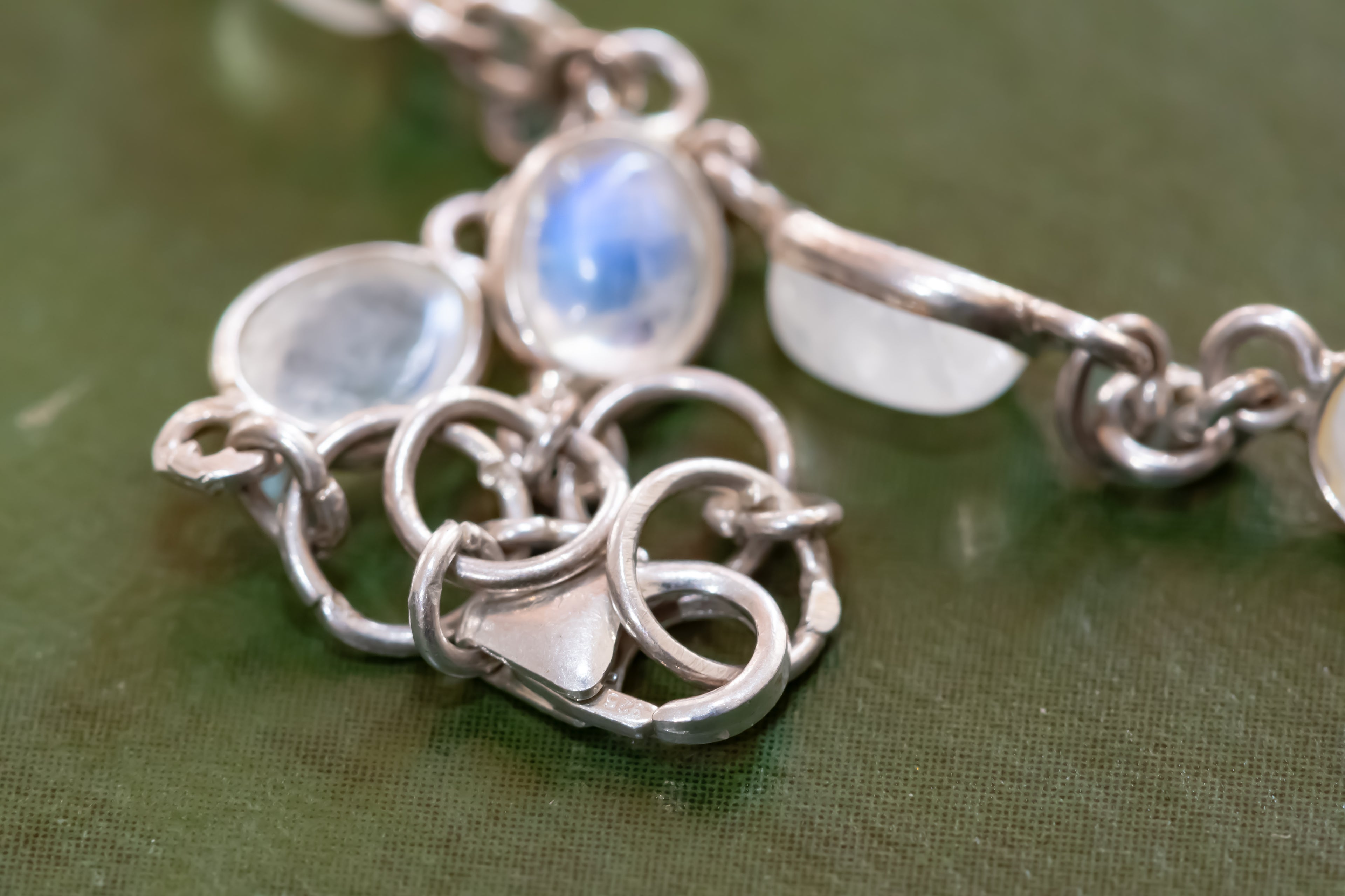 A close-up of a silver-toned chain bracelet featuring several oval moonstone gemstones with blue and white iridescent qualities, resting on a textured olive green surface.