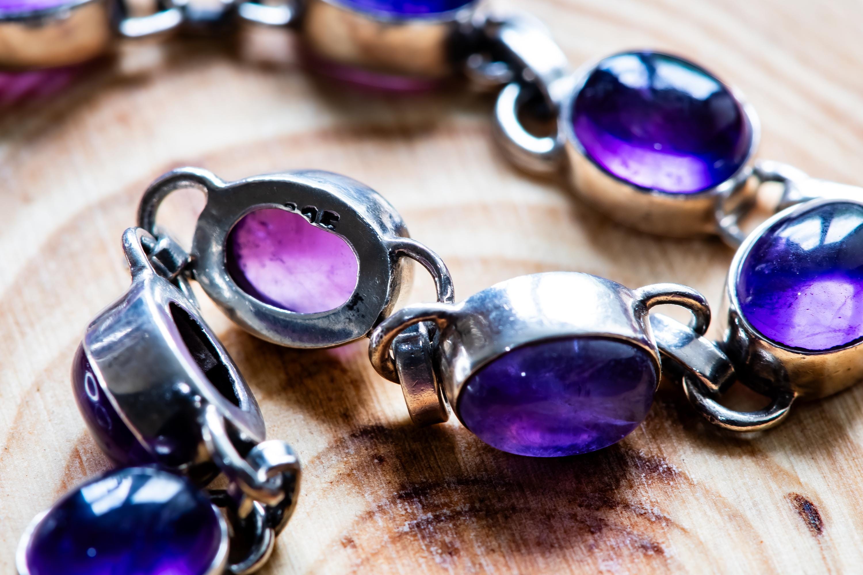 A silver link bracelet featuring multiple oval-shaped, deep purple amethyst cabochons set in polished metal bezels. Each individual link is connected by small silver loops, and one of the metal settings is clearly stamped with 925.
