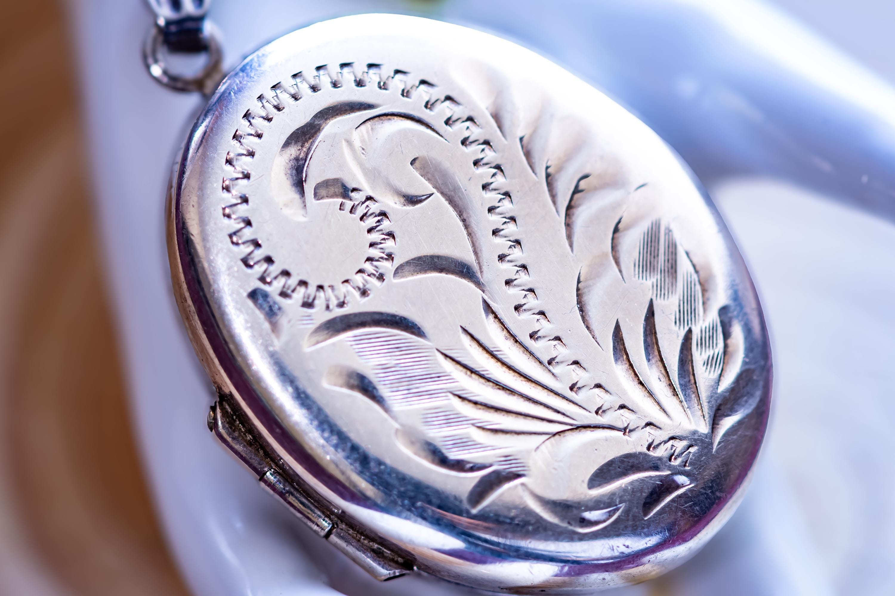 An oval silver locket pendant featuring an intricate engraved floral pattern on its front surface. The metal has a polished, reflective finish with deep etched lines creating a swirling leaf design. A small metal loop is attached at the top.