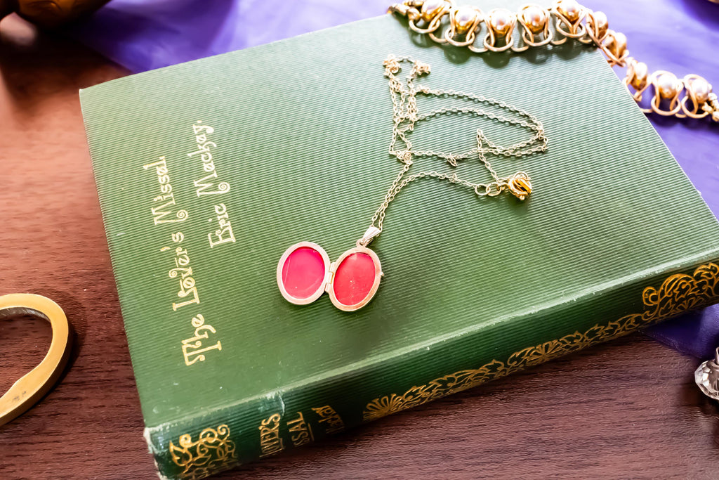 A gold-toned metal locket necklace with an open oval pendant revealing bright pink interiors, resting on a green hardcover book titled The Lover's Missal by Eric Mackay, featuring gold embossed lettering and decorative border details.