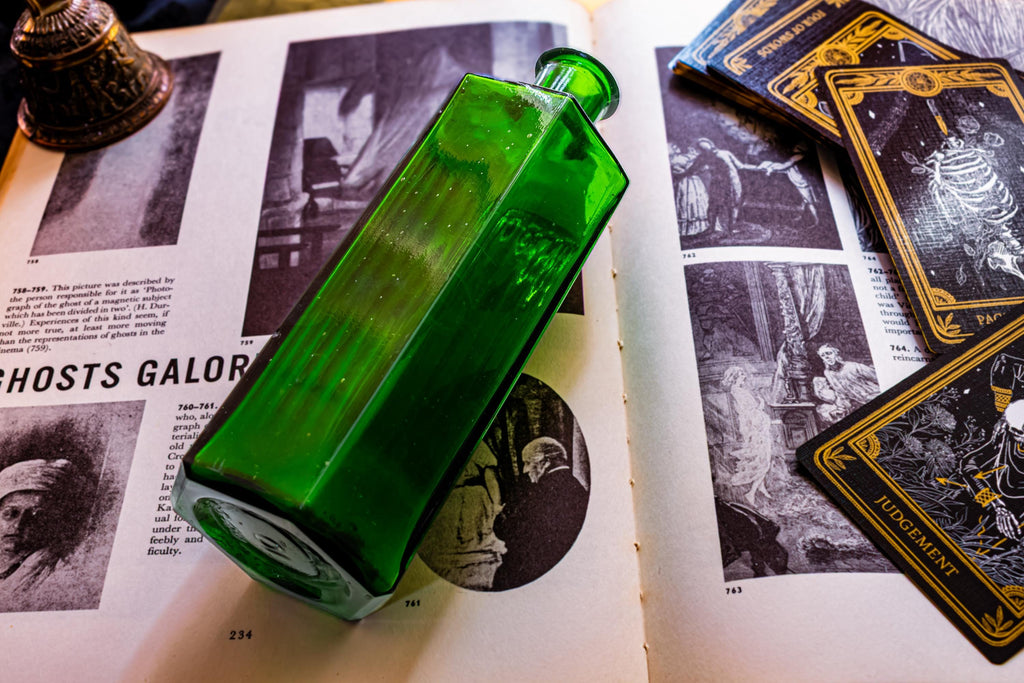 A rectangular emerald green glass bottle with a narrow neck, positioned diagonally across an open book featuring black and white illustrations, text titled GHOSTS GALORE, and several black and gold tarot cards nearby.