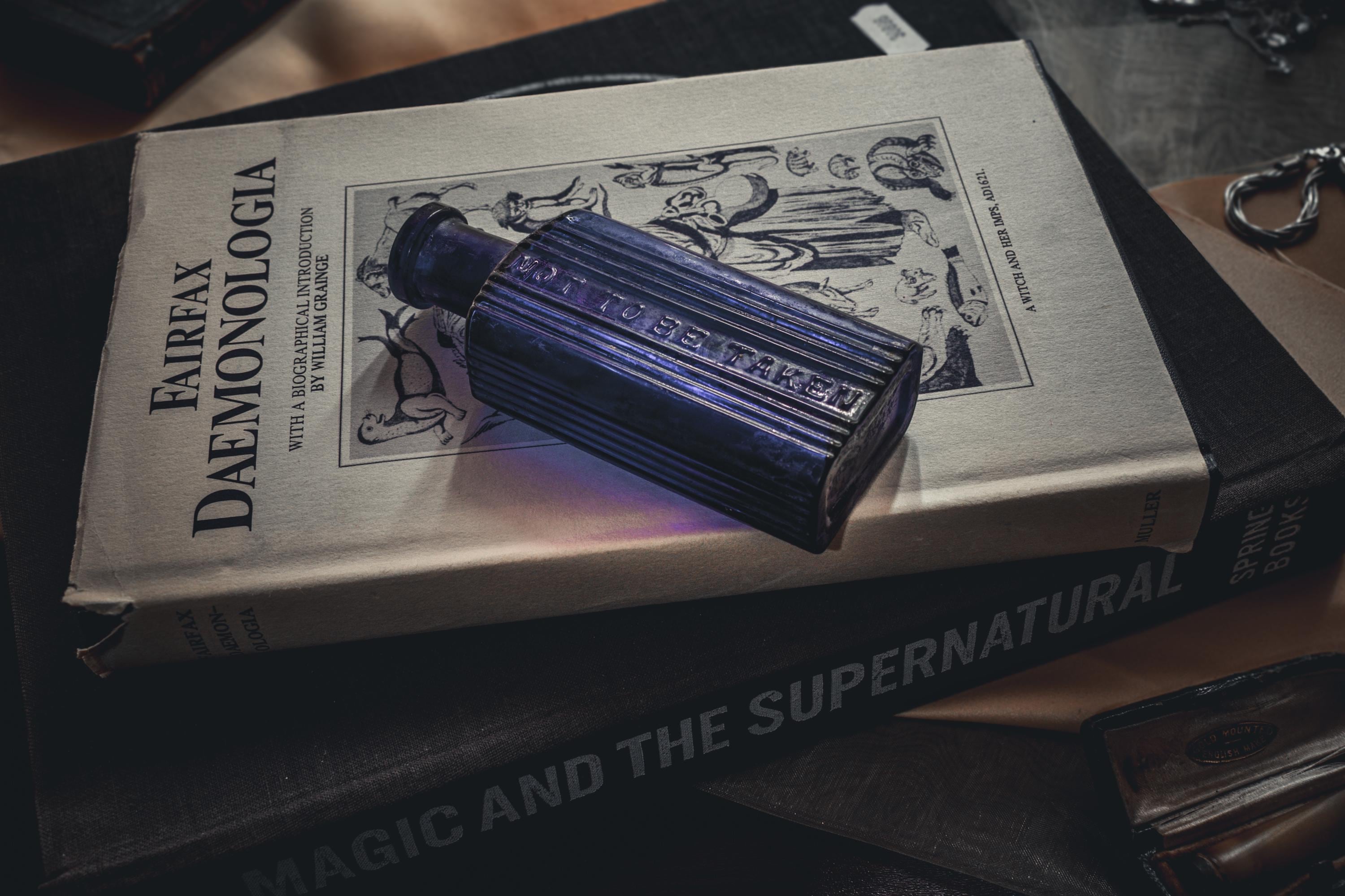 A dark blue rectangular glass bottle with ribbed sides and the embossed text NOT TO BE TAKEN, resting on a book titled FAIRFAX DAEMONOLOGIA, which is placed atop another book titled MAGIC AND THE SUPERNATURAL.