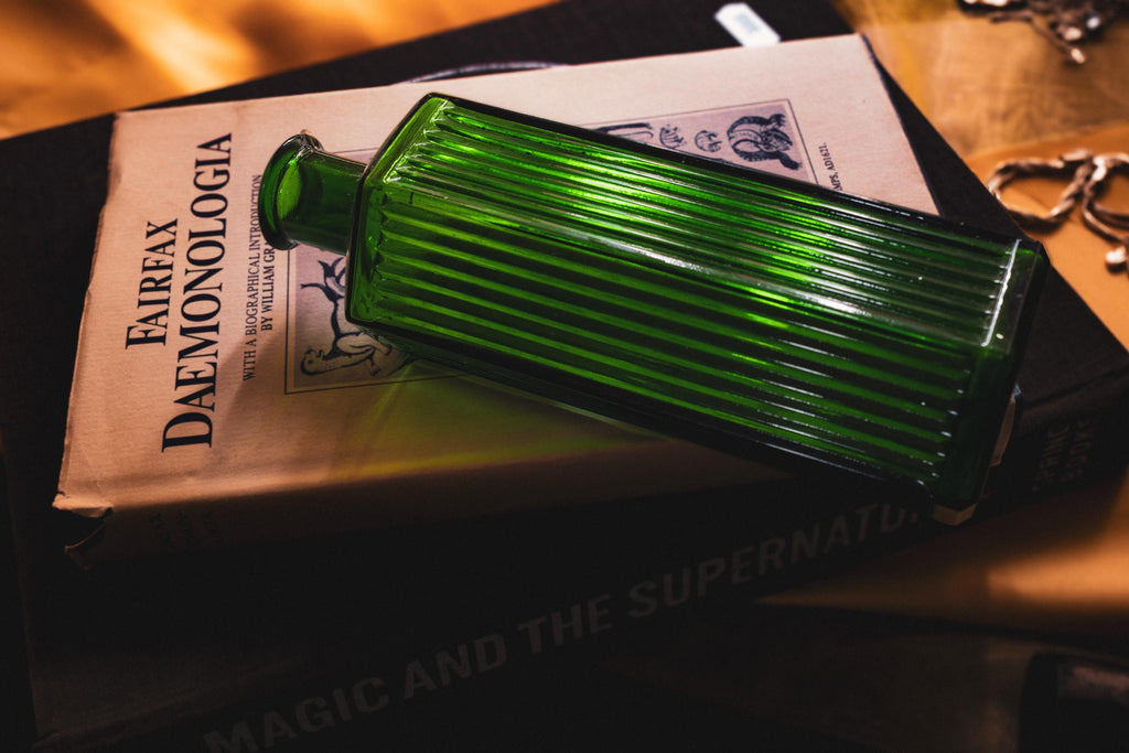 A rectangular, emerald green glass bottle with vertical ribbed detailing along its sides. The bottle is positioned horizontally across a book titled Fairfax Daemonologia, which features a black and white illustration.