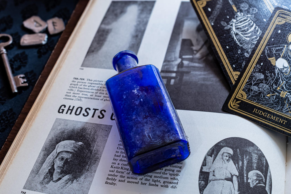 A rectangular cobalt blue glass bottle resting on an open book titled Ghosts. The book features black and white illustrations of figures and text. Nearby are a metal key, small stone runes, and a tarot card labelled Judgement.