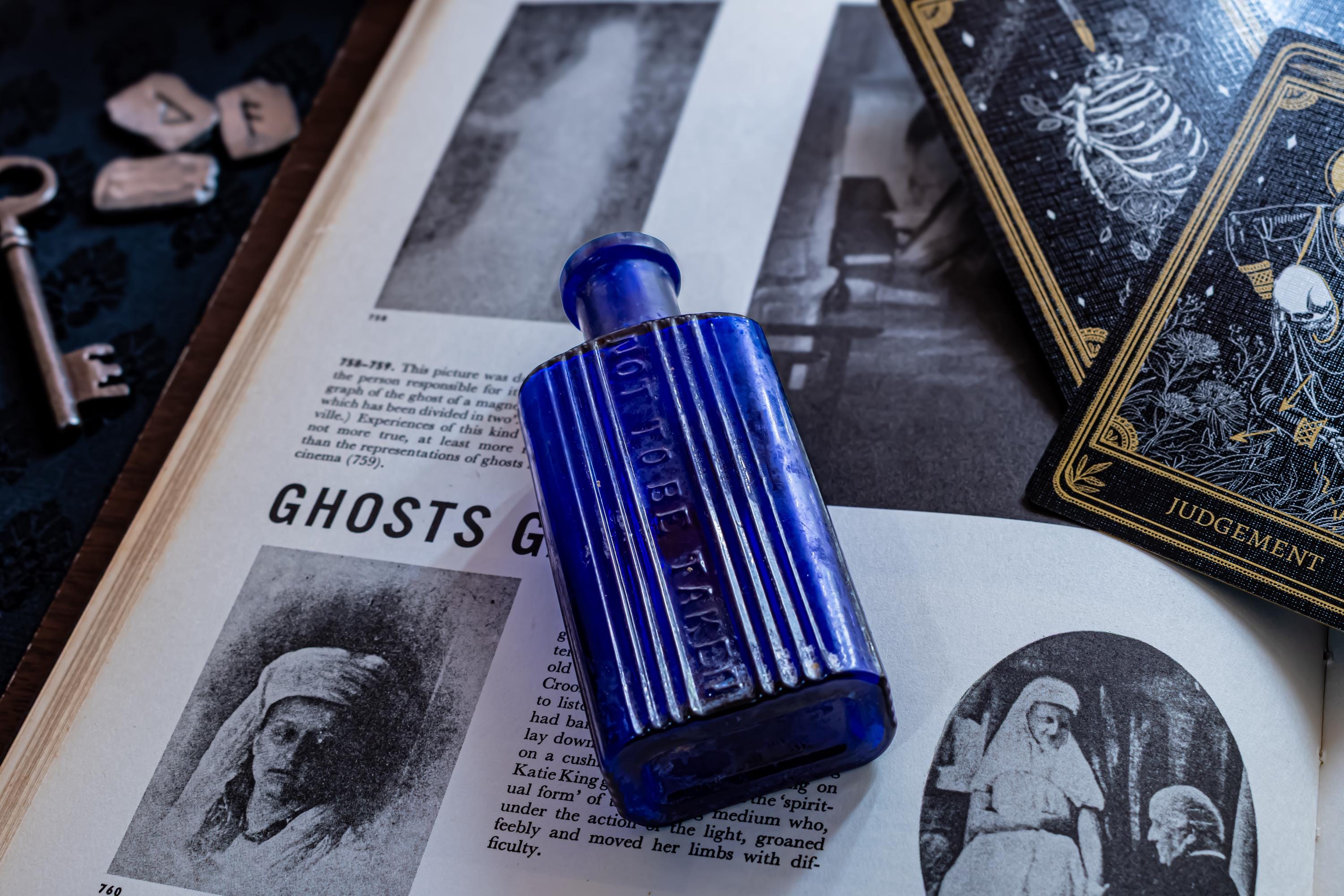 A rectangular cobalt blue glass bottle with vertical ribbing and the embossed text NOT TO BE TAKEN on the front. The bottle is positioned on an open book page featuring black and white illustrations and text about ghosts.