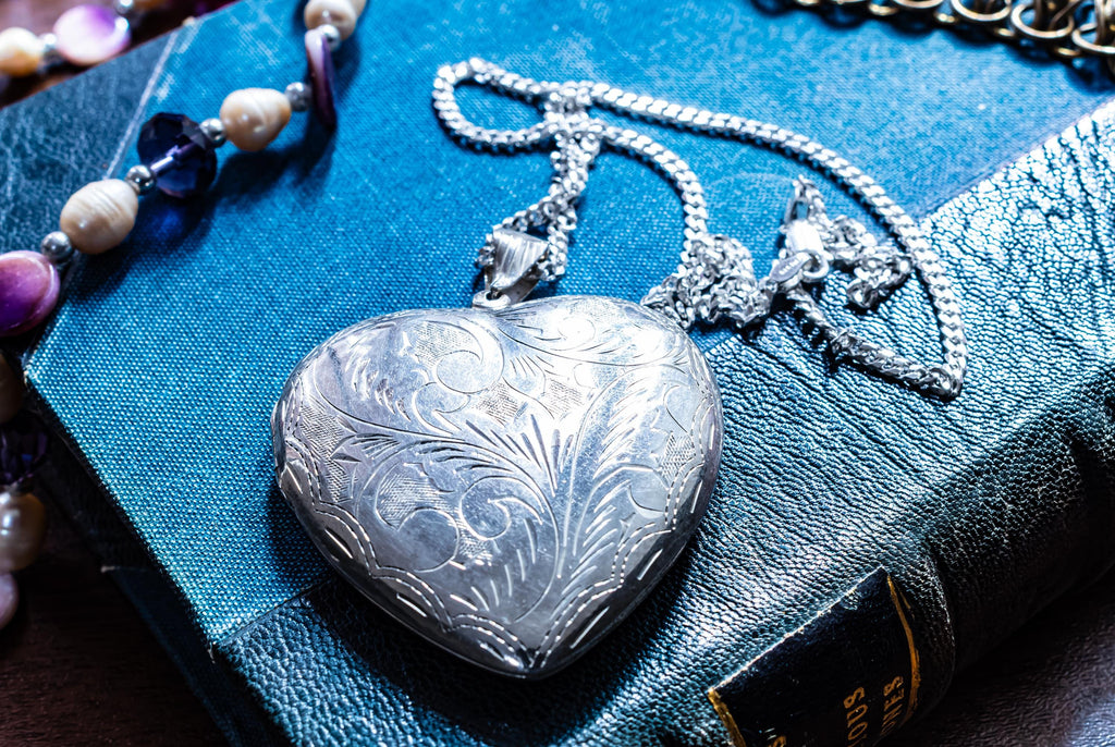 A silver-toned heart-shaped vintage locket pendant featuring intricate floral engravings on the front surface, suspended from a matching silver-toned metal link chain that rests on a textured blue book cover with visible gold lettering.