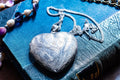 A silver-toned heart-shaped vintage locket pendant featuring intricate floral engravings on the front surface, suspended from a matching silver-toned metal link chain that rests on a textured blue book cover with visible gold lettering.