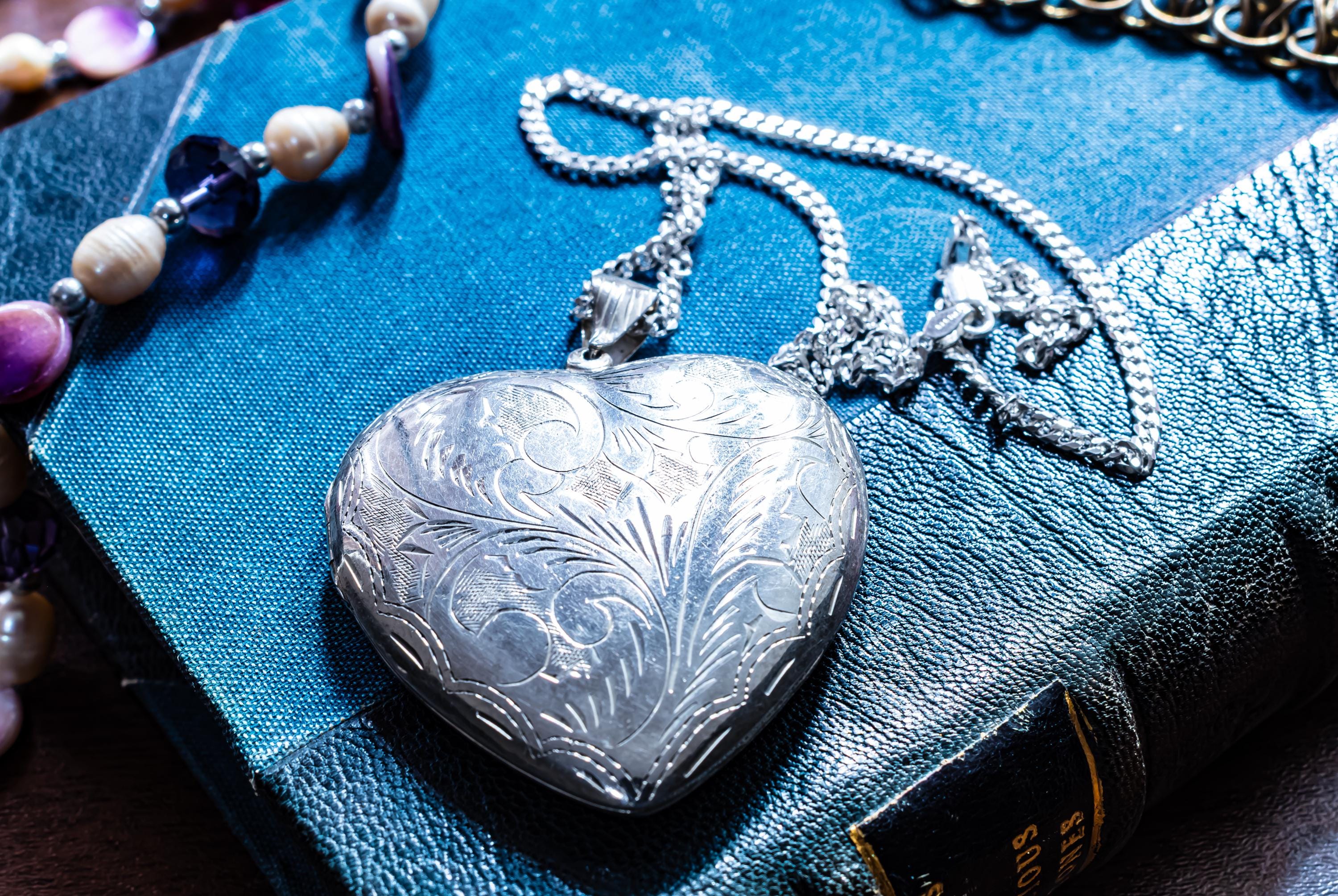 A silver-toned heart-shaped vintage locket pendant featuring intricate floral engravings on the front surface, suspended from a matching silver-toned metal link chain that rests on a textured blue book cover with visible gold lettering.