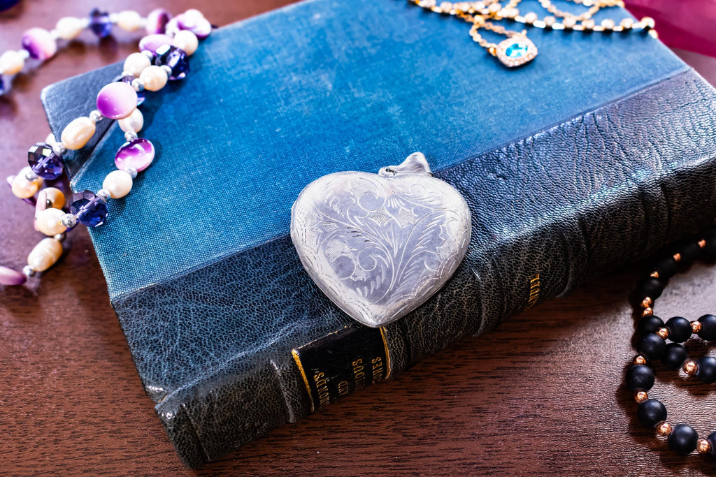 A silver-toned heart-shaped vintage locket featuring an intricate engraved floral pattern on its surface. The locket is positioned on top of a textured blue book, surrounded by various beaded necklaces and a gold-toned chain with a blue stone.