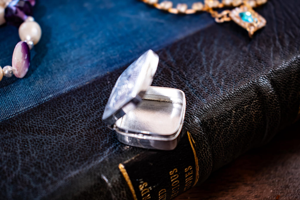 An image of an opened Tiny square antique silver pill box with engraved floral patterns sitting on a dark textured book