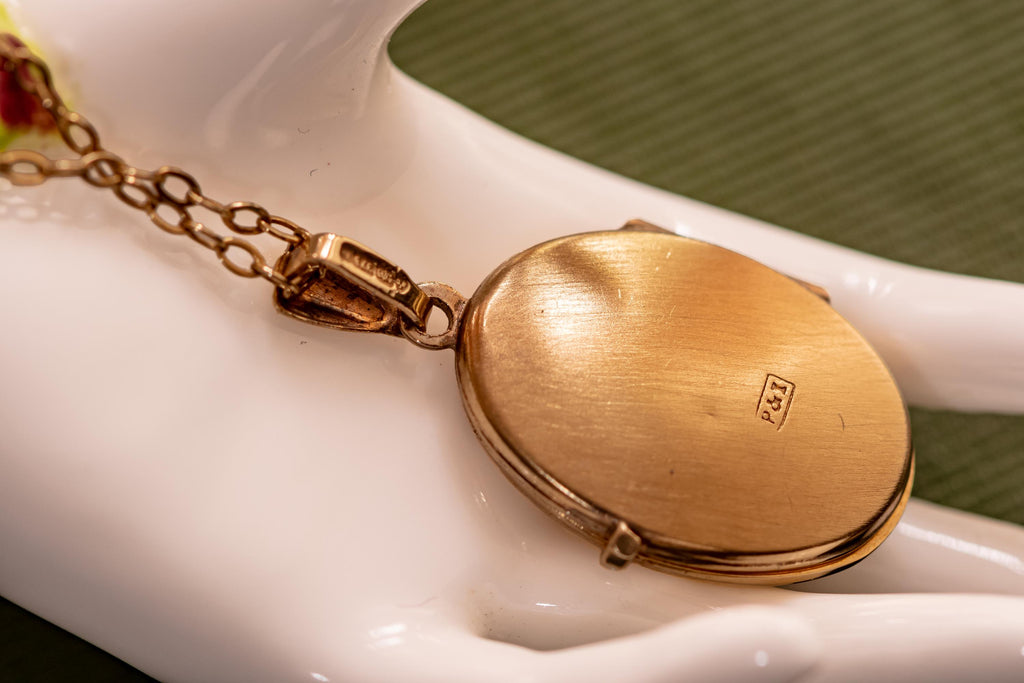 An oval gold-toned locket pendant attached to a delicate link chain. The back of the locket features a smooth, brushed metallic finish with a small rectangular stamp containing the letters P&I clearly visible on the surface.