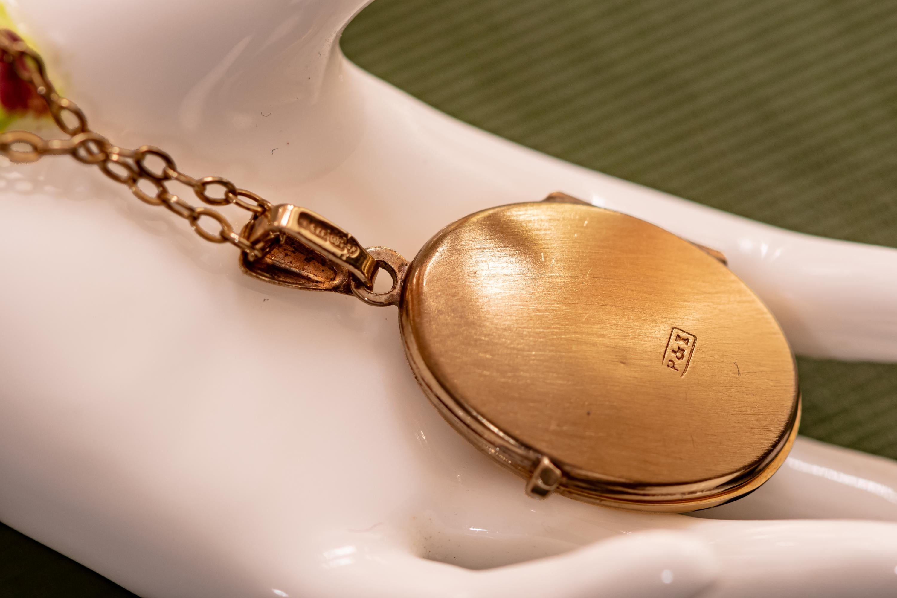 An oval gold-toned locket pendant attached to a delicate link chain. The back of the locket features a smooth, brushed metallic finish with a small rectangular stamp containing the letters P&I clearly visible on the surface.