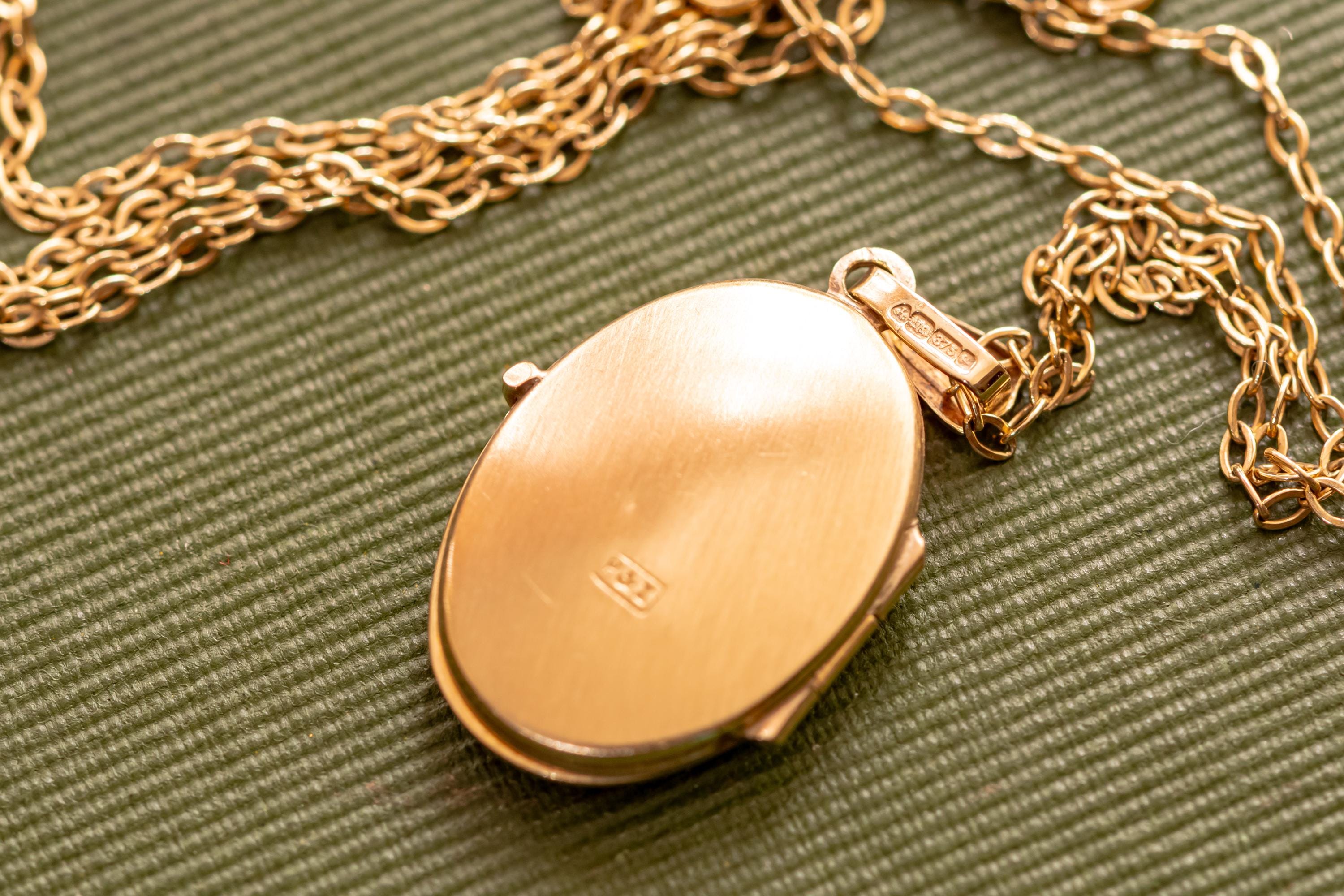 An oval-shaped rose gold locket pendant with a smooth, polished finish, suspended from a matching delicate rose gold chain. The locket features a small, rectangular hallmark stamp on its surface and a bail with visible engraved markings.