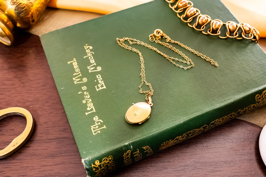 A gold oval locket on a delicate chain resting on a green hardcover book titled The Lover's Missal by Eric Mackay. The book features gold embossed lettering on the cover and spine, accompanied by a gold link bracelet nearby.