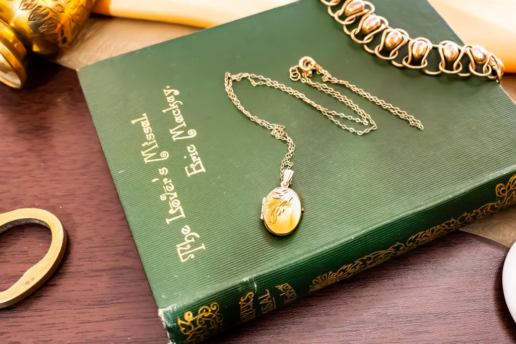 A gold oval locket on a delicate chain resting on a green hardcover book titled The Lover's Missal by Eric Mackay. The locket features an engraved feather design on its front surface. A gold-toned bracelet is visible in the corner.