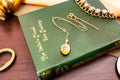 A gold oval locket on a delicate chain resting on a green hardcover book titled The Lover's Missal by Eric Mackay. The locket features an engraved feather design on its front surface. A gold-toned bracelet is visible in the corner.