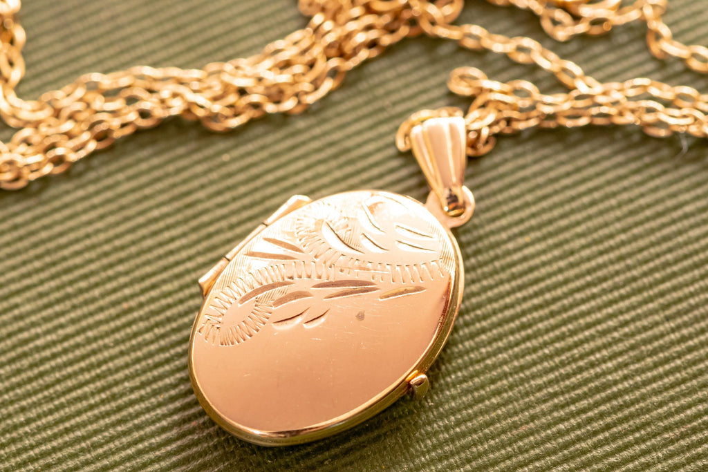 An oval-shaped gold-toned locket pendant suspended from a matching gold-toned link chain. The front surface of the locket features an engraved decorative pattern consisting of curved lines and small hatch marks across the top half.