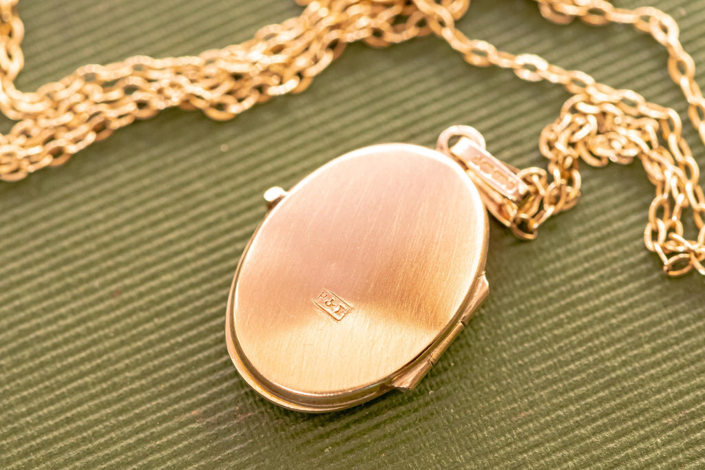 A gold-toned oval locket pendant suspended from a matching link chain. The locket features a brushed metal finish on its front surface with a small rectangular hallmark stamped in the centre and a bail at the top for the chain.